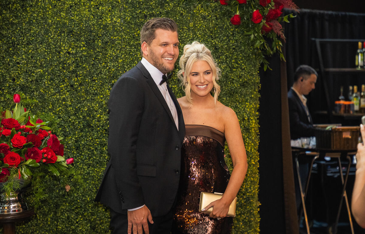 Smiling couple in formal attire posing against a green hedge wall with red rose bouquets in the foreground and background.