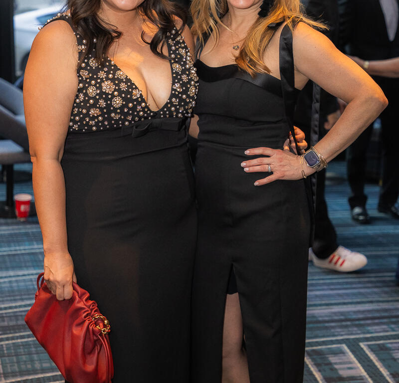 Two women in black evening gowns smile and pose together at a formal event; one holds a red clutch bag.