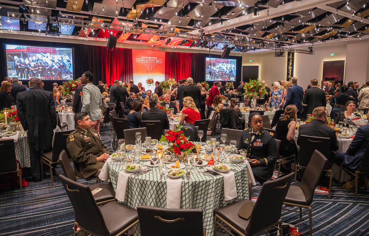 Banquet hall full of guests in formal attire, round tables with red floral centerpieces, stage with red curtains behind.