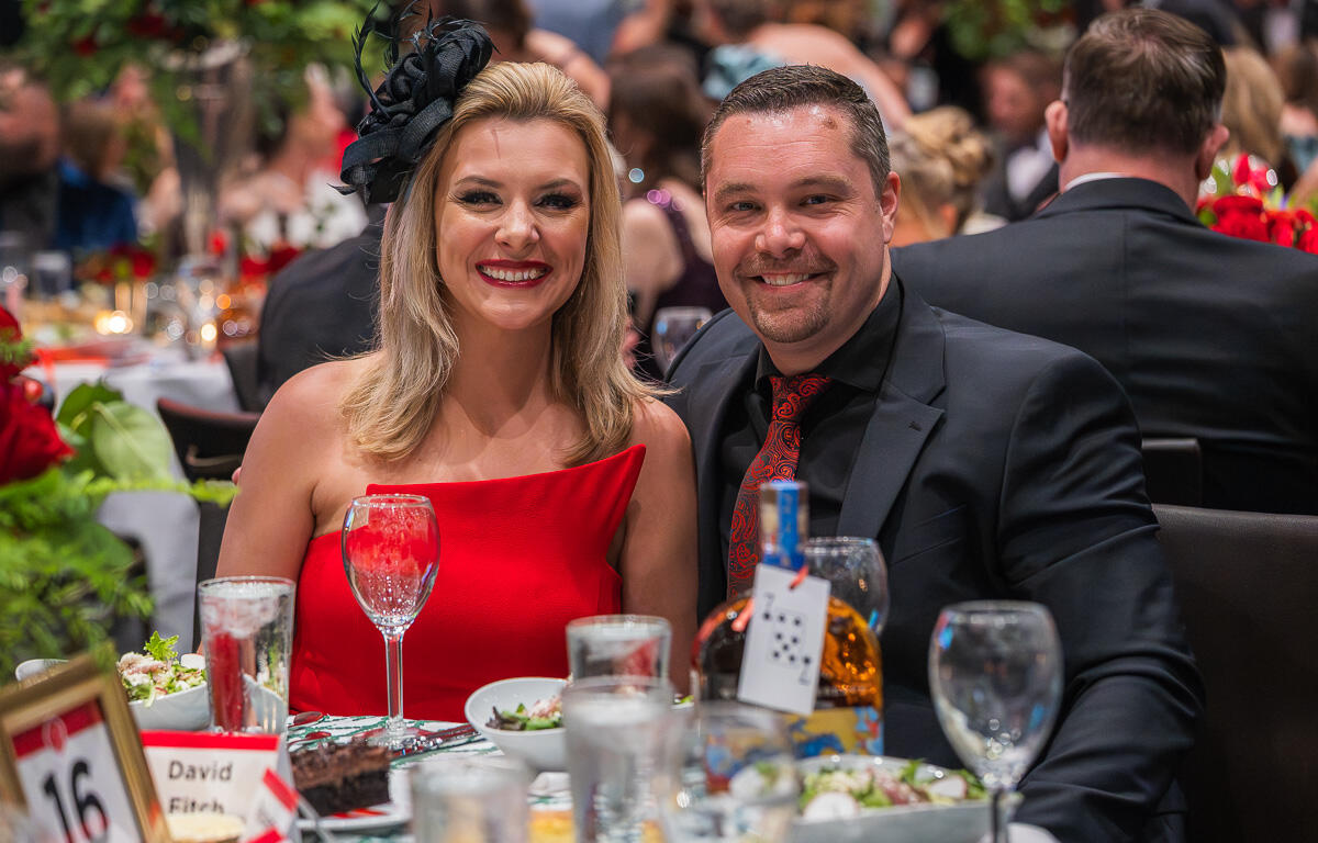 Smiling couple seated at a banquet table during a formal event; woman wears a red dress and a black fascinator, man in a dark suit with a red tie.