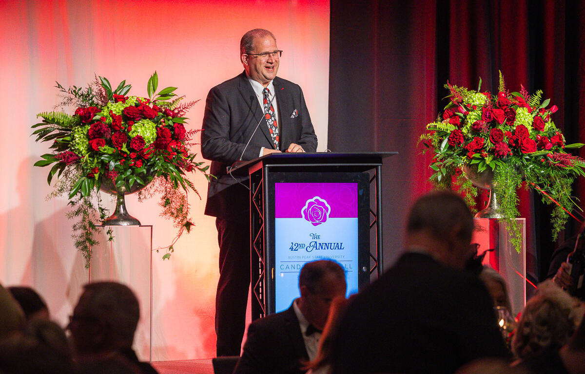 Speaker at a lectern delivering a speech at a formal event, with large red flower arrangements on stage.