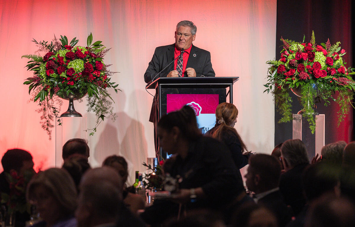 Man in a suit delivering a speech at a podium with red backdrop and large floral arrangements on each side.