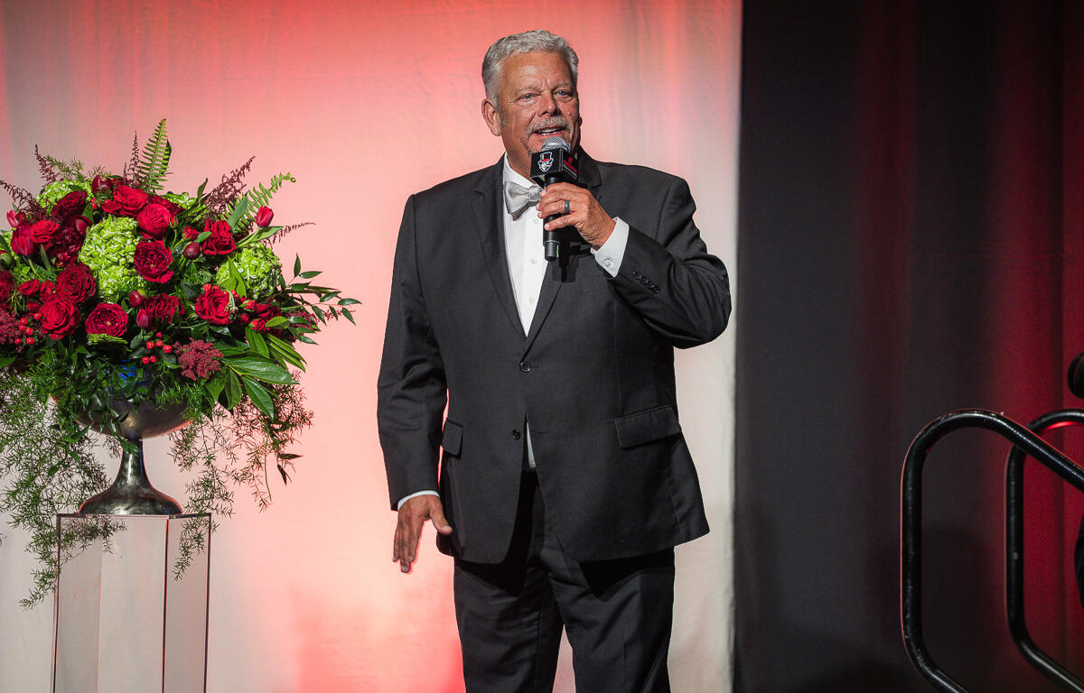 Older man in a black tuxedo and white bow tie speaks into a microphone on a stage with a large red flower arrangement beside him on a pedestal