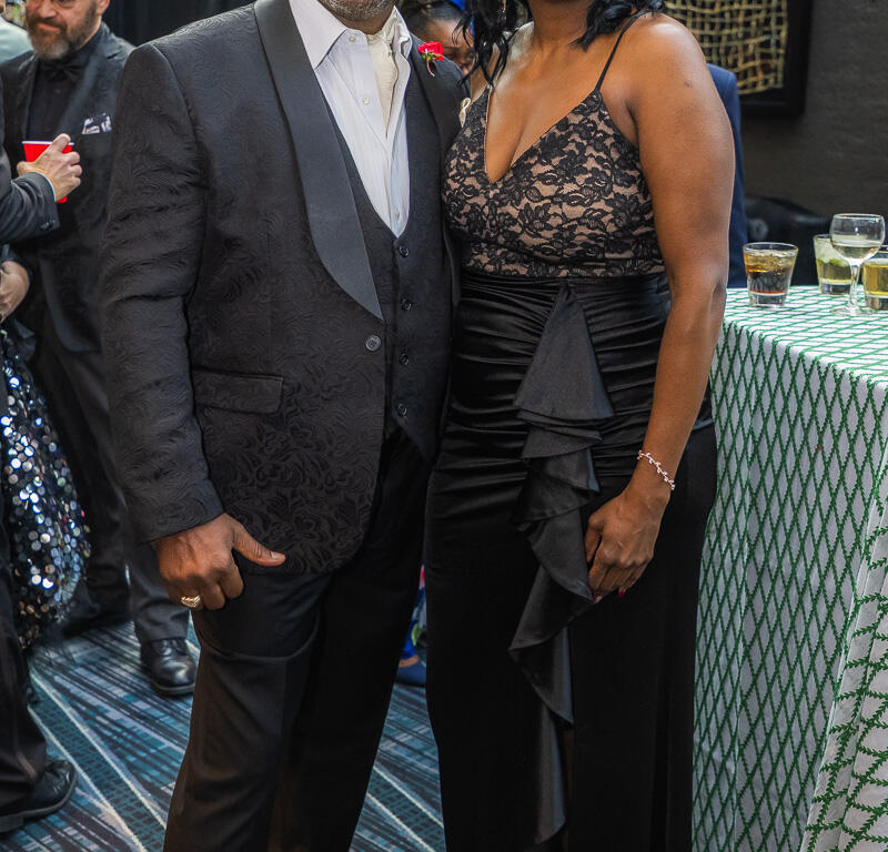 Smiling couple in formal attire posing at a reception; man in a dark patterned suit and vest, woman in a black lace gown.