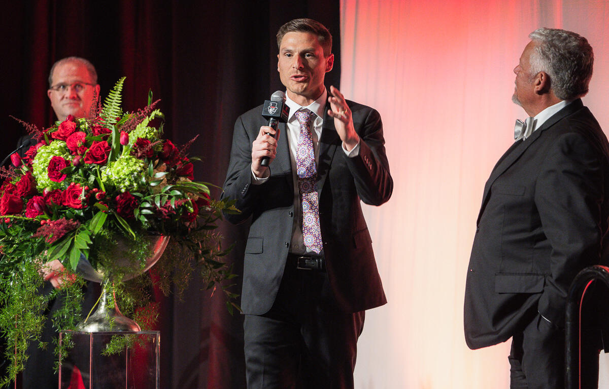 Man in a dark suit speaks into a microphone on a stage, with two other men in suits nearby and a large red floral arrangement on a pedestal to the left.