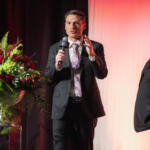 Man in a dark suit speaks into a microphone on a stage, with two other men in suits nearby and a large red floral arrangement on a pedestal to the left.