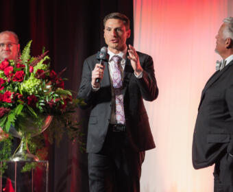 Man in a dark suit speaks into a microphone on a stage, with two other men in suits nearby and a large red floral arrangement on a pedestal to the left.