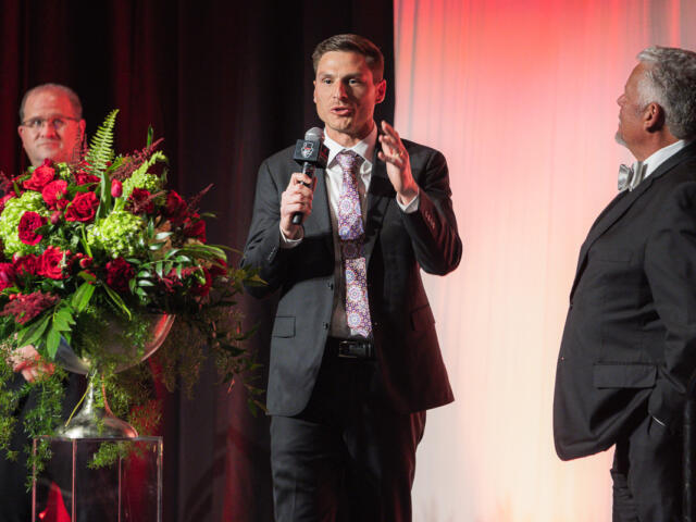 Man in a dark suit speaks into a microphone on a stage, with two other men in suits nearby and a large red floral arrangement on a pedestal to the left.