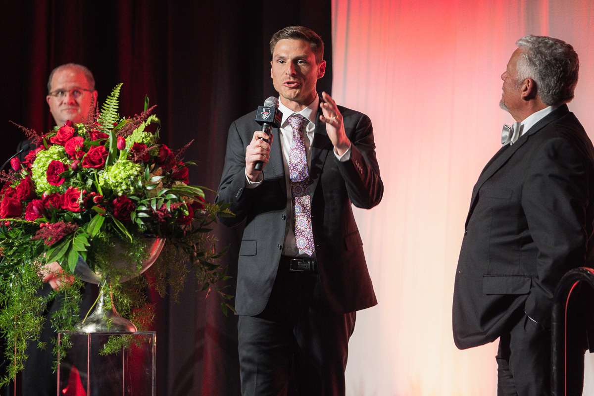 Man in a dark suit speaks into a microphone on a stage, with two other men in suits nearby and a large red floral arrangement on a pedestal to the left.
