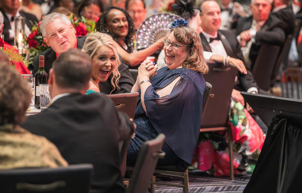 Two women in evening gowns laugh at a formal gala table, with wine glasses and a floral centerpiece nearby.