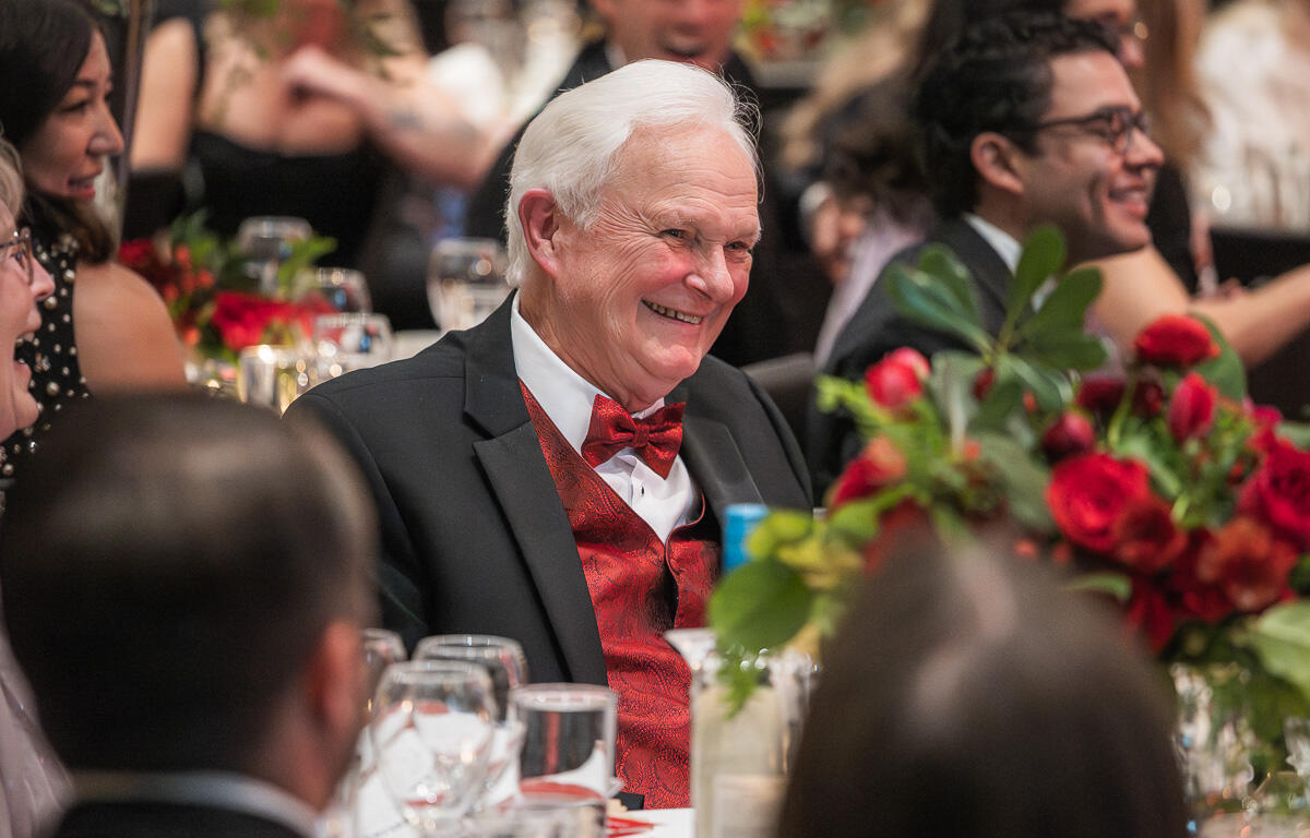 Smiling elderly man in a black suit with a red vest and bow tie at a banquet table with a red floral centerpiece in the foreground.