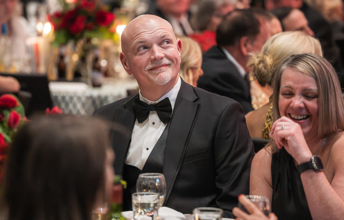 Man in a black tuxedo and bow tie smiles as he looks up at a formal dinner table, a laughing woman beside him fades into the scene.