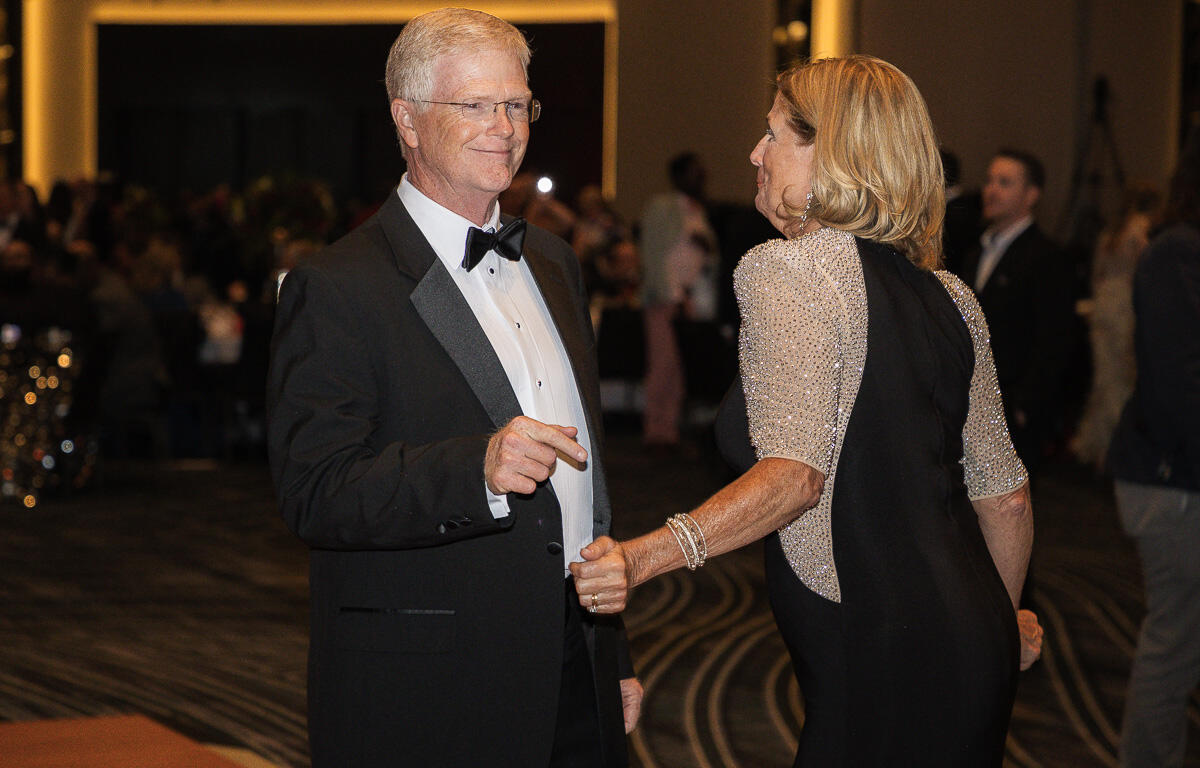 Older man in a tuxedo and a woman in a beaded gown dance and smile at a formal gala venue.