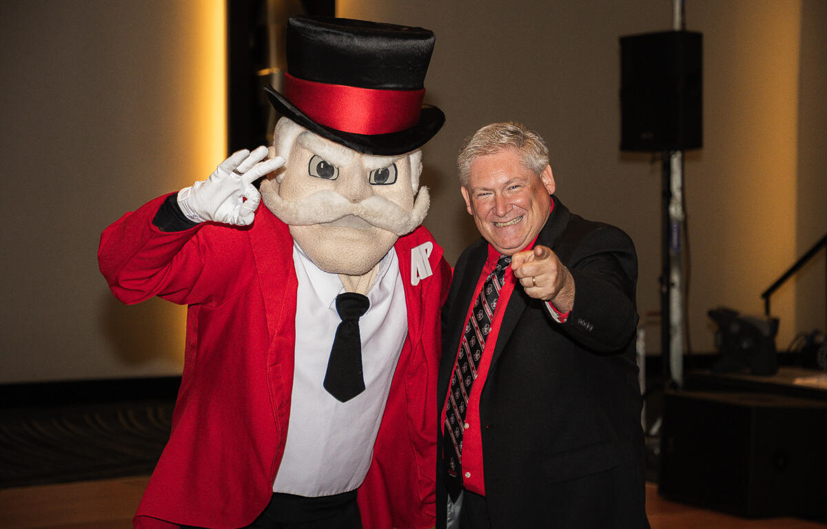Man in a black suit and red tie points at the camera, posing with a large cartoon mascot in a red jacket and top hat at an indoor event.