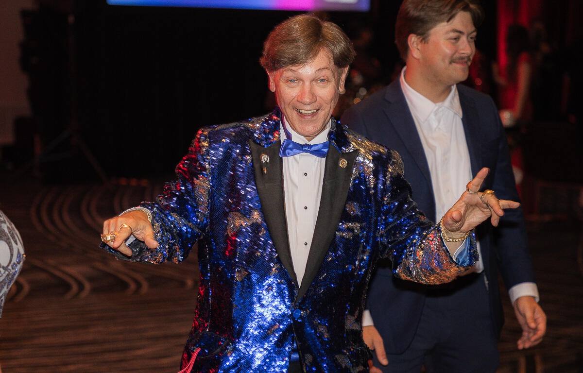 Smiling man in a blue sequined tuxedo dancing at a party, with another man in a suit in the background.