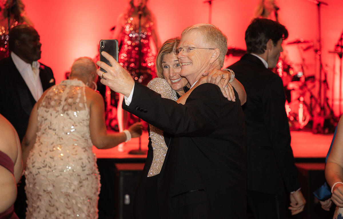 Smiling couple in formal wear taking a selfie on a dance floor, with a red-lit stage and band in the background.