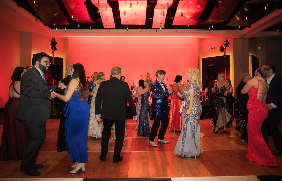 Guests in formal evening wear dancing on a wooden floor at a gala with a red-lit stage and musicians in the background.