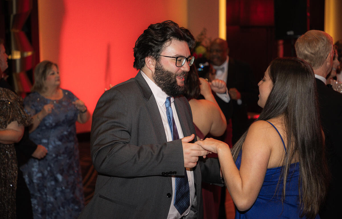 Bearded man in a gray suit dancing with a woman in a blue dress at a formal party, warm red lighting in the background.