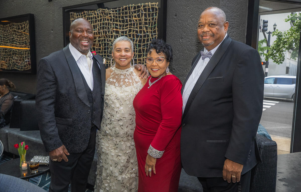 Group of four adults in formal wear posing for a photo at an indoor event, smiling.