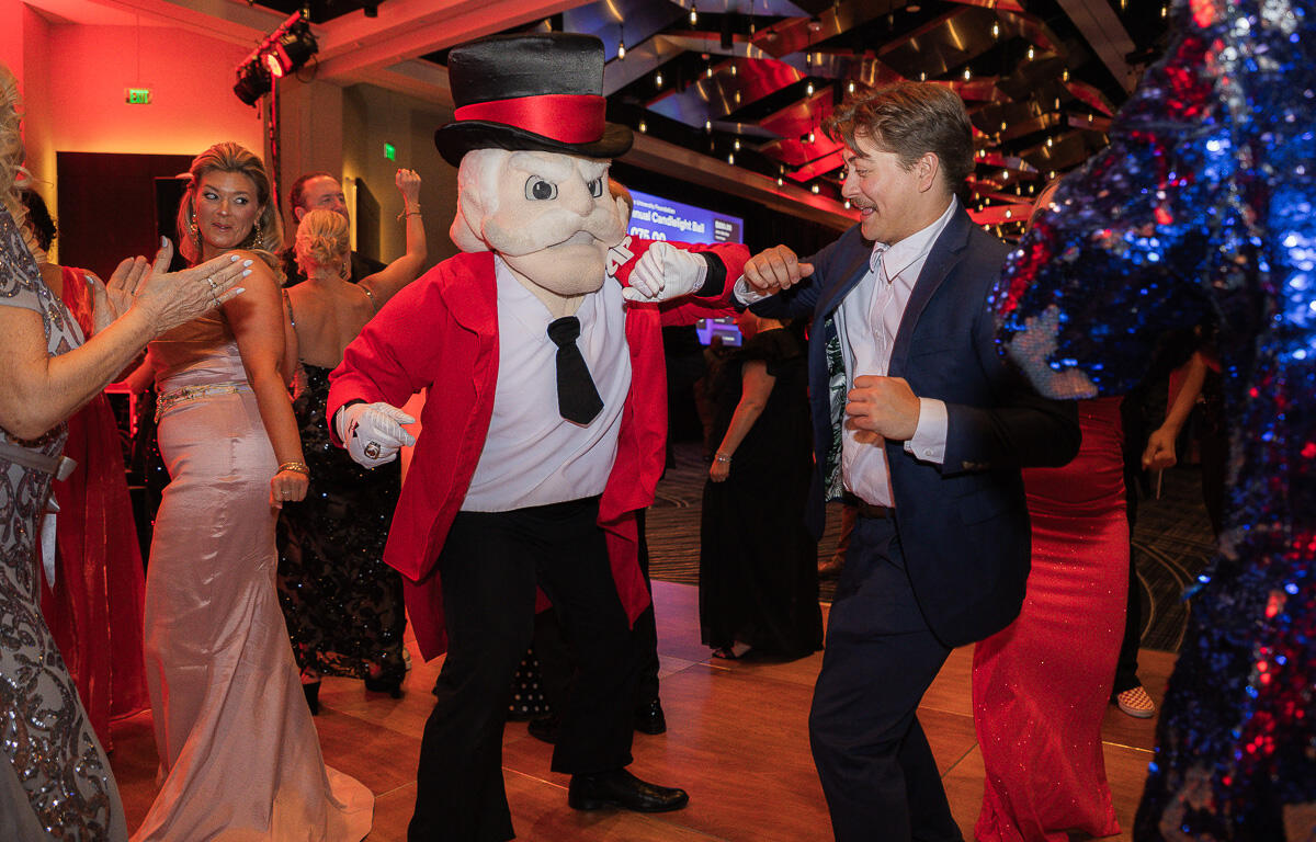 Man in a navy suit dances with a cartoonish mascot in a red jacket and top hat at a lively party.
