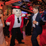 Man in a navy suit dances with a cartoonish mascot in a red jacket and top hat at a lively party.