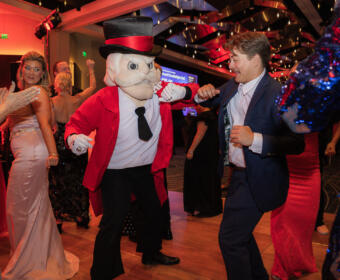 Man in a navy suit dances with a cartoonish mascot in a red jacket and top hat at a lively party.
