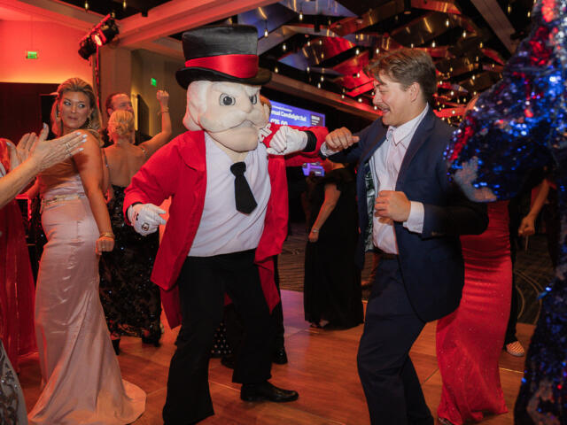 Man in a navy suit dances with a cartoonish mascot in a red jacket and top hat at a lively party.