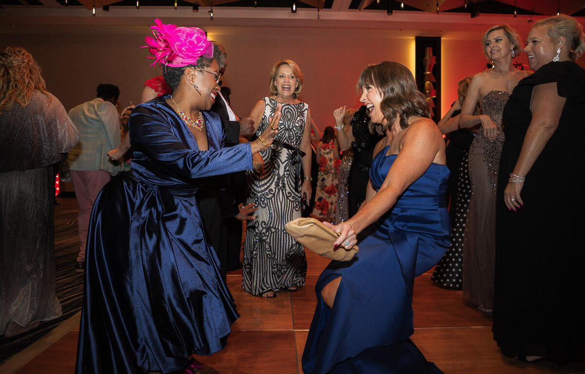 Two women in formal gowns dance on a lit dance floor, high-fiving and smiling at a lively party.