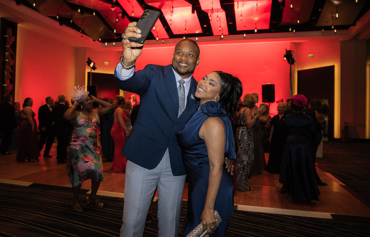Couple in formal attire posing for a selfie on a crowded dance floor at a gala event with guests in evening wear in the background.