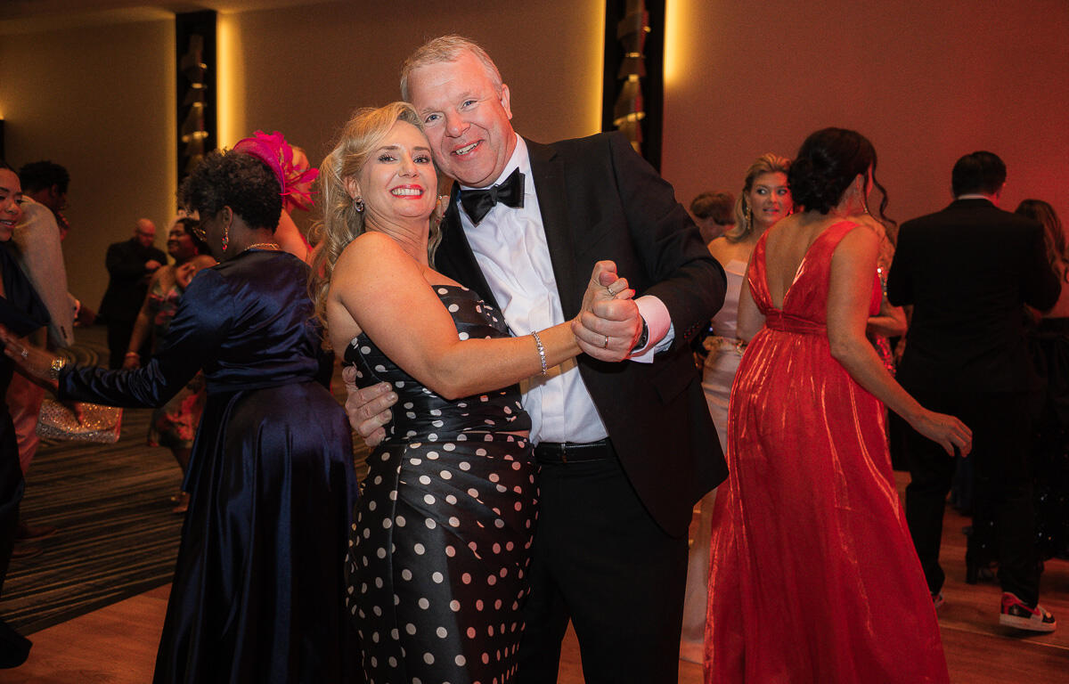 Couple dancing closely at a formal party, the woman in a black polka-dot dress and the man in a tuxedo smile at the camera while dancing with others in the background.