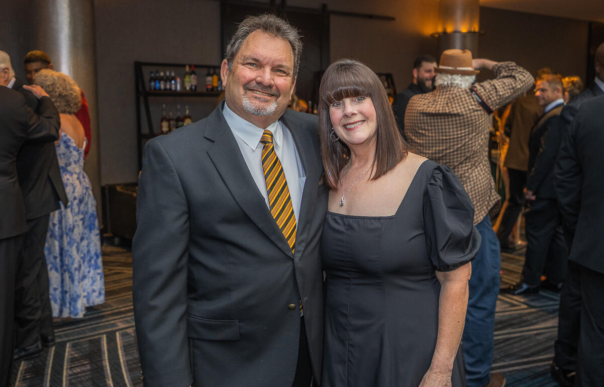 A smiling couple in formal attire posing together at a social event, with a bar area and other guests in the background.