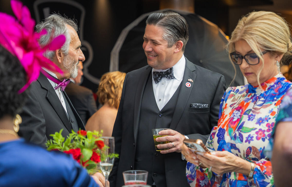 Smiling man in a black suit and bow tie chats with an older gentleman at a cocktail party with drinks nearby.