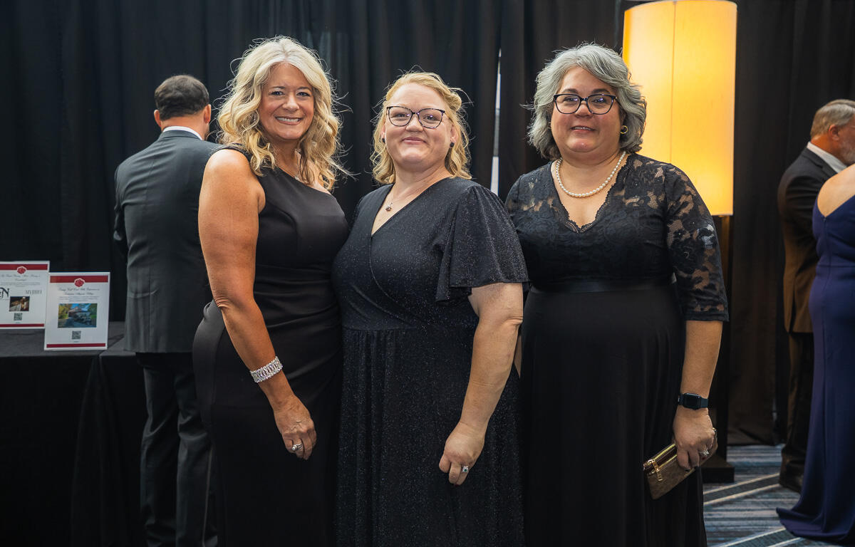 Three women in black formal dresses pose and smile at a gala, standing together indoors near a warm floor lamp and dark backdrop.