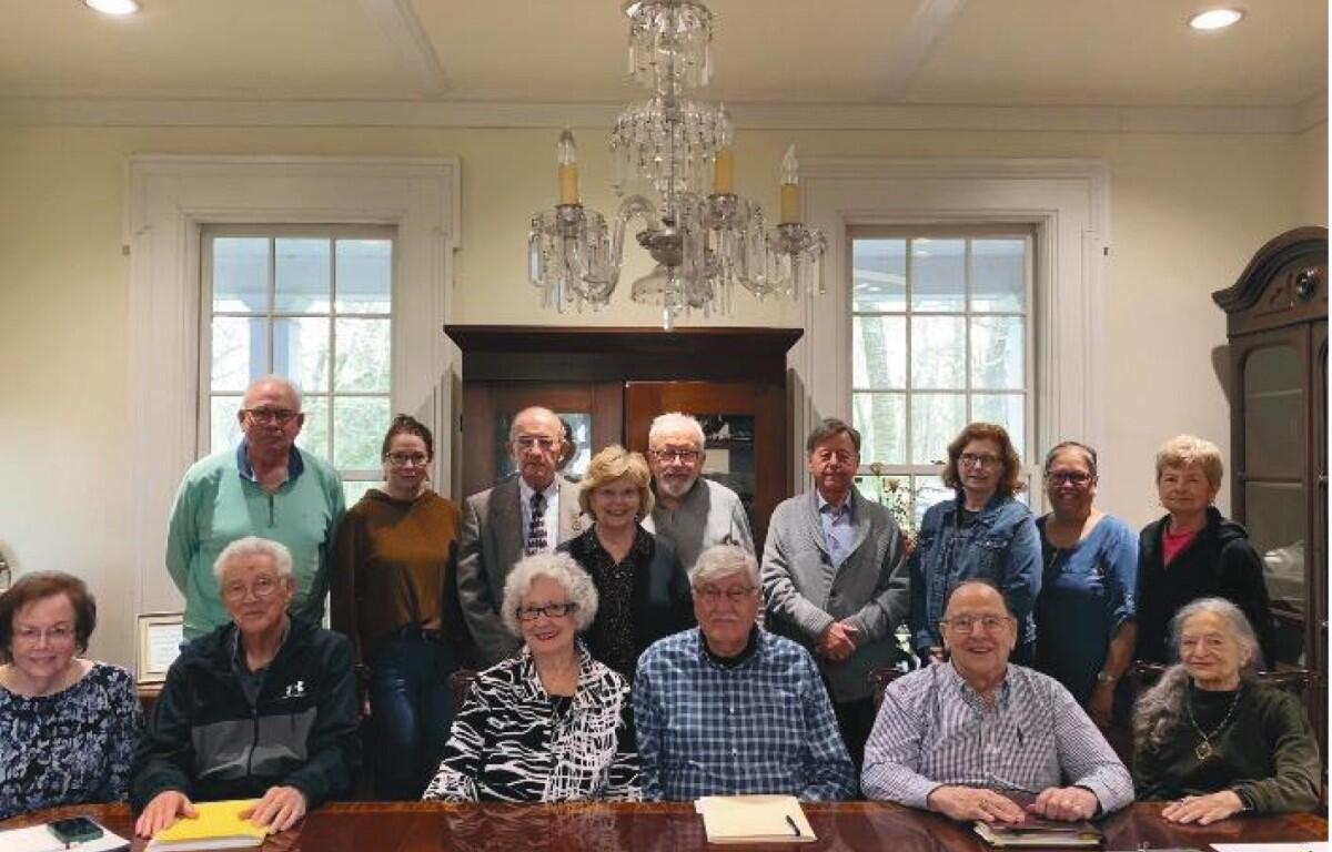 Group of seniors and adults posed for a group photo around a large wooden conference table in a bright room with a chandelier overhead.