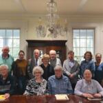 Group of seniors and adults posed for a group photo around a large wooden conference table in a bright room with a chandelier overhead.
