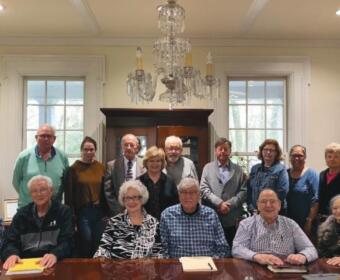 Group of seniors and adults posed for a group photo around a large wooden conference table in a bright room with a chandelier overhead.