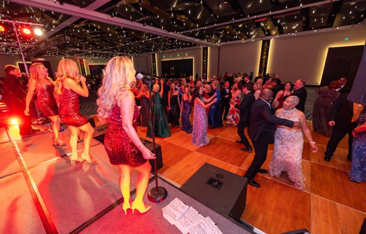 Singer in a red sequin dress performs on stage as guests in formal attire dance on a wooden floor beneath string lights in a ballroom event.