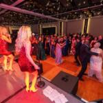 Singer in a red sequin dress performs on stage as guests in formal attire dance on a wooden floor beneath string lights in a ballroom event.