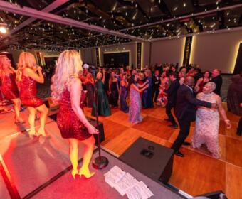 Singer in a red sequin dress performs on stage as guests in formal attire dance on a wooden floor beneath string lights in a ballroom event.