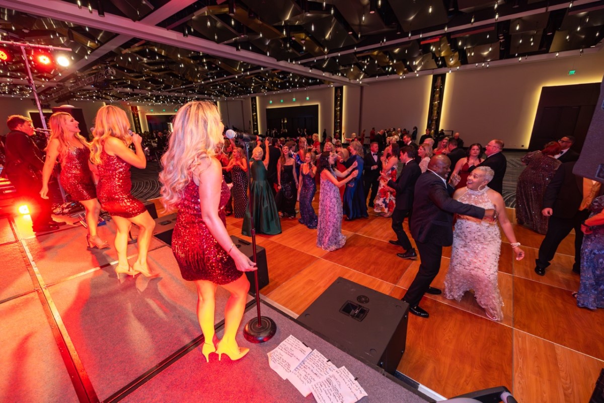Singer in a red sequin dress performs on stage as guests in formal attire dance on a wooden floor beneath string lights in a ballroom event.
