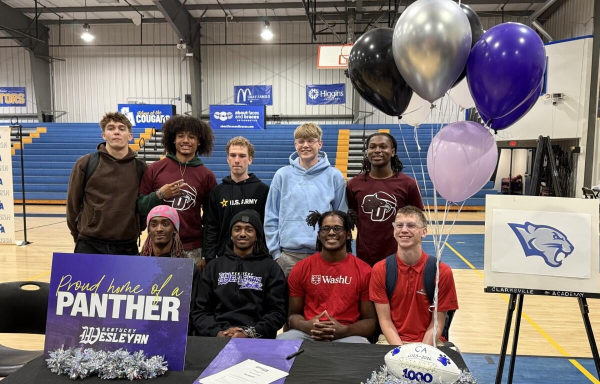 Group of ten students posing in a school gym for a photo, with a purple 'Proud home of a Panther' sign and silver/purple balloons at a signing/event table in front of bleachers.