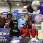 Group of ten students posing in a school gym for a photo, with a purple 'Proud home of a Panther' sign and silver/purple balloons at a signing/event table in front of bleachers.