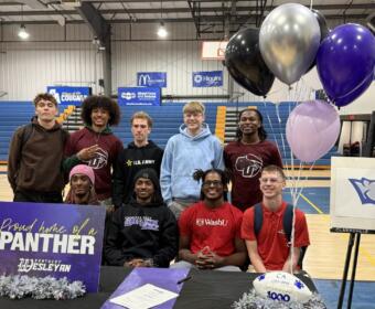 Group of ten students posing in a school gym for a photo, with a purple 'Proud home of a Panther' sign and silver/purple balloons at a signing/event table in front of bleachers.