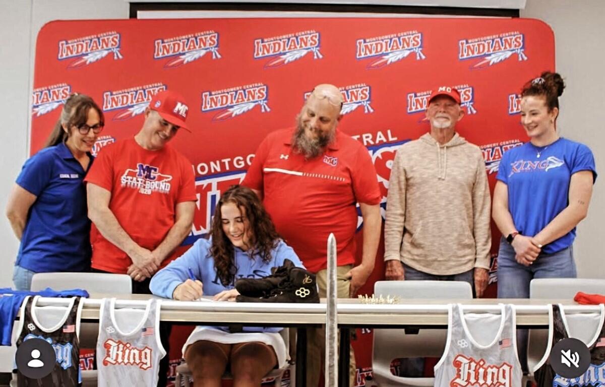 Woman seated at a table signs a jersey while five teammates watch in front of a red team backdrop.