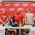 Woman seated at a table signs a jersey while five teammates watch in front of a red team backdrop.