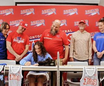 Woman seated at a table signs a jersey while five teammates watch in front of a red team backdrop.