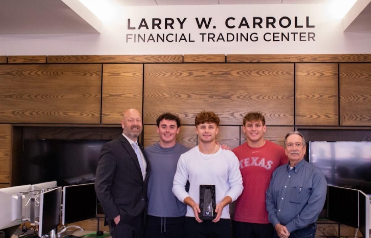 Five men pose in a modern office under a sign that reads LARRY W. CARROLL FINANCIAL TRADING CENTER; the center man holds a black trophy or award.