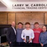 Five men pose in a modern office under a sign that reads LARRY W. CARROLL FINANCIAL TRADING CENTER; the center man holds a black trophy or award.
