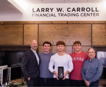 Five men pose in a modern office under a sign that reads LARRY W. CARROLL FINANCIAL TRADING CENTER; the center man holds a black trophy or award.