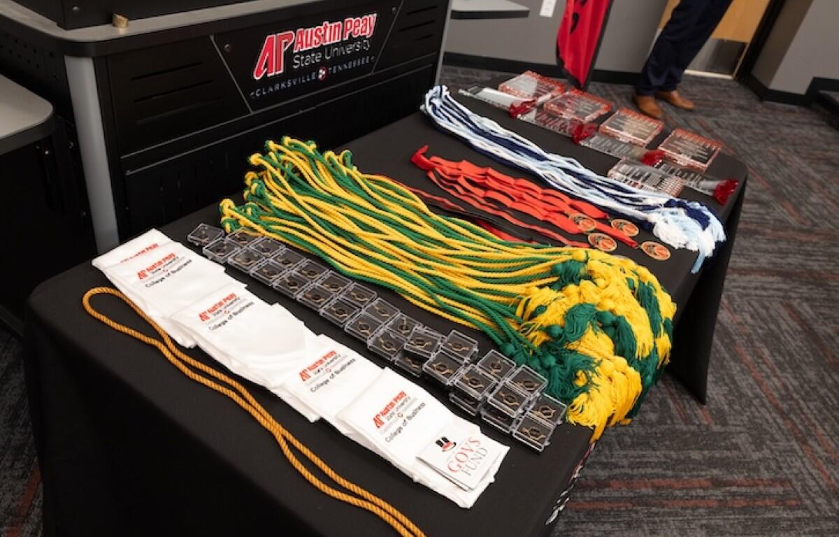 Table with award medals on green and yellow cords, red/white/blue lanyards, and white drawstring bags at a university event with branding in the background.